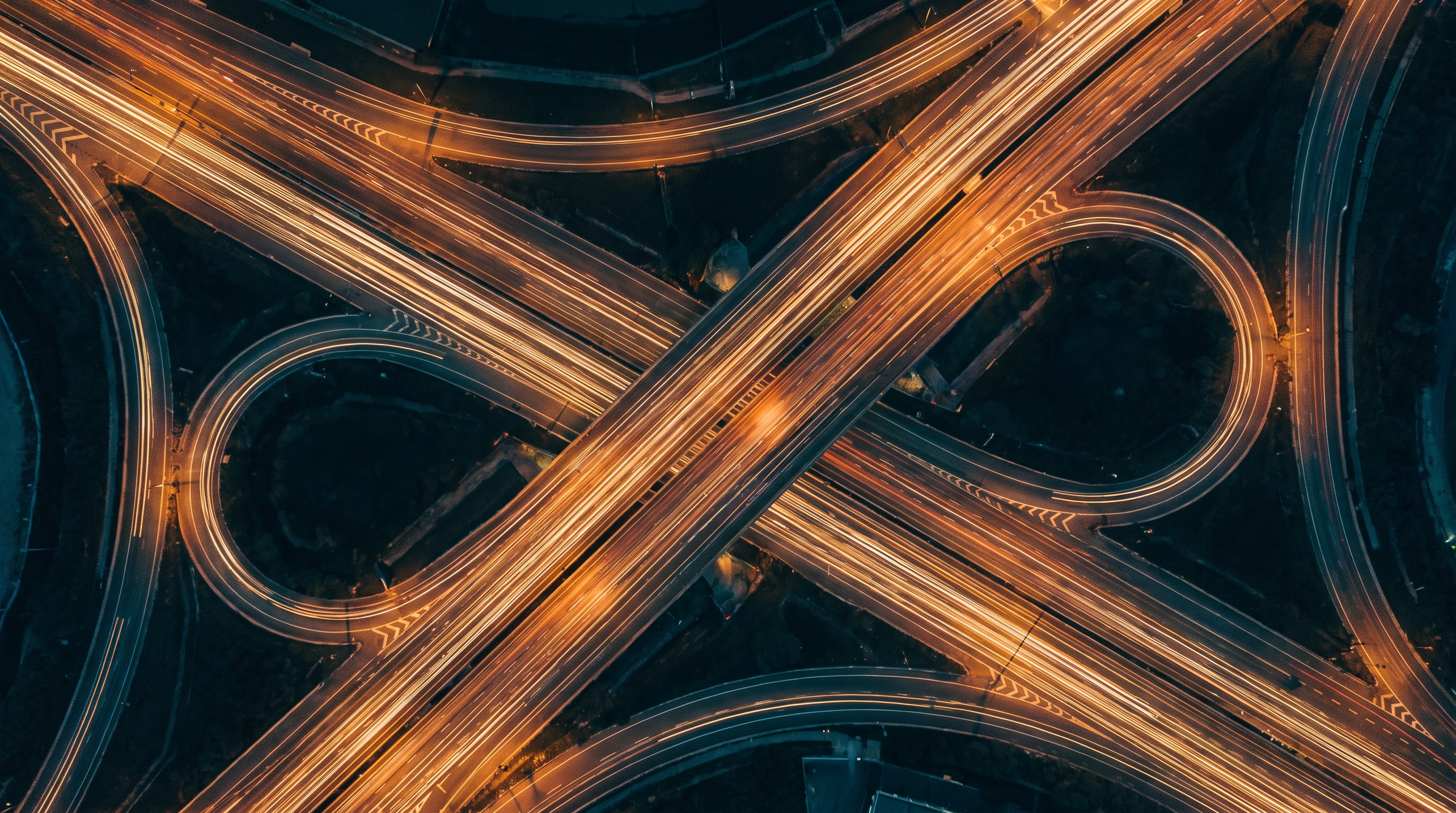 Long-exposure aerial of a highway interchange at night, amber light-trails braiding through deep teal darkness